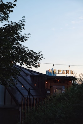 A rooftop venue with a sign in Cyrillic script, partially obscured by foliage. The structure features a sloped roof and string lights hanging overhead. The sky is clear and softly lit during sunset, casting a serene ambiance.