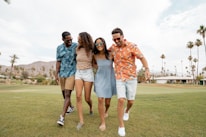 Four friends are walking together outdoors on a grassy area. They are smiling and dressed in casual, colorful summer clothing. The sky is partly cloudy, and there are palm trees and mountains in the background, suggesting a warm, sunny location.