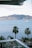 A serene coastal view with a calm ocean and a sailboat docked in the water. In the foreground, several palm trees are standing along a beach area with multiple sun loungers arranged neatly. The background displays a range of green, lush hills or mountains under a light blue sky with sparse clouds.
