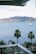 A serene coastal view with a calm ocean and a sailboat docked in the water. In the foreground, several palm trees are standing along a beach area with multiple sun loungers arranged neatly. The background displays a range of green, lush hills or mountains under a light blue sky with sparse clouds.