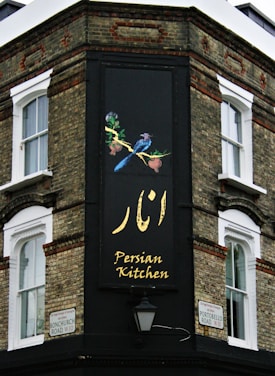 The corner of a brick building features a large, vertical sign with elegant writing in both English and another script, reading 'Persian Kitchen'. Above the text, an artistic illustration shows two birds perched on a branch with pomegranates. The building is located at the intersection of Bonchurch Road and Portobello Road.