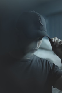 A person is partially obscured by shadows, wearing a dark cap and t-shirt. The ambiance is moody and dimly lit, with soft focus lending an air of mystery.