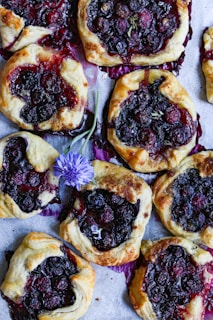 A collection of rustic, open-faced pastries filled with dark, juicy berries on a baking sheet. The pastries have a golden-brown flaky crust, with deep purple and red berries peeking through the center. A delicate purple flower lies among the pastries, adding a splash of color to the arrangement.