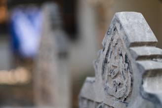 A close-up of a weathered stone carving with intricate details, possibly part of a gravestone or monument, against a blurred background.