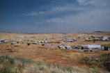 A sprawling landscape dotted with numerous tents and makeshift shelters, spread across a dry, grassy terrain under an expansive, partly cloudy sky. The settlement appears temporary and scattered across the arid landscape, suggesting it might be a refugee camp or a similar setup.