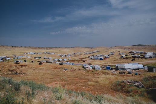 A sprawling landscape dotted with numerous tents and makeshift shelters, spread across a dry, grassy terrain under an expansive, partly cloudy sky. The settlement appears temporary and scattered across the arid landscape, suggesting it might be a refugee camp or a similar setup.