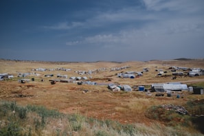 A sprawling landscape dotted with numerous tents and makeshift shelters, spread across a dry, grassy terrain under an expansive, partly cloudy sky. The settlement appears temporary and scattered across the arid landscape, suggesting it might be a refugee camp or a similar setup.