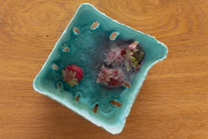 A blue cardboard container holds strawberries with visible mold growth. The once fresh strawberries are now covered in gray and white mold, giving them a decaying appearance. The container rests on a wooden surface with a grainy texture.