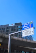 A tall road sign is situated in front of a modern office building under a clear blue sky. The sign indicates directions to various locations, including Constanța and Ploiești. Below, concrete infrastructure suggests an urban environment.