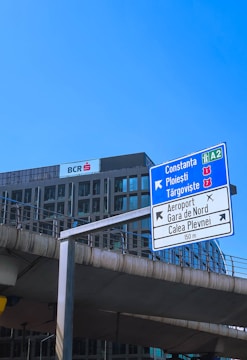 A tall road sign is situated in front of a modern office building under a clear blue sky. The sign indicates directions to various locations, including Constanța and Ploiești. Below, concrete infrastructure suggests an urban environment.