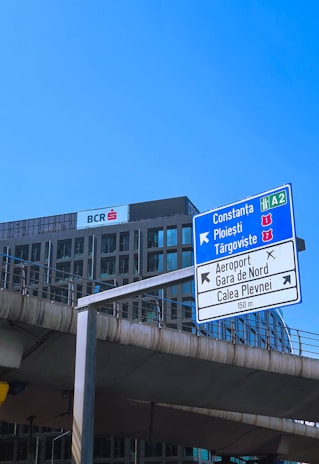 A tall road sign is situated in front of a modern office building under a clear blue sky. The sign indicates directions to various locations, including Constanța and Ploiești. Below, concrete infrastructure suggests an urban environment.
