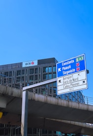 A tall road sign is situated in front of a modern office building under a clear blue sky. The sign indicates directions to various locations, including Constanța and Ploiești. Below, concrete infrastructure suggests an urban environment.