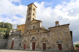 A historic stone church with a tall bell tower stands under a partly cloudy sky. The architecture features ornate designs and multiple entrances with wooden doors. Surrounding the church are lush green trees and shrubbery.