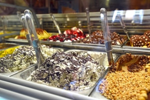 A display of various gelato flavors, with metal scoops positioned in each container. The gelato includes flavors with chocolate, nuts, and fruit toppings visible. The setting appears to be a gelato shop with multiple trays arranged in a row.
