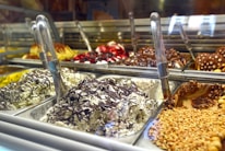 A display of various gelato flavors, with metal scoops positioned in each container. The gelato includes flavors with chocolate, nuts, and fruit toppings visible. The setting appears to be a gelato shop with multiple trays arranged in a row.