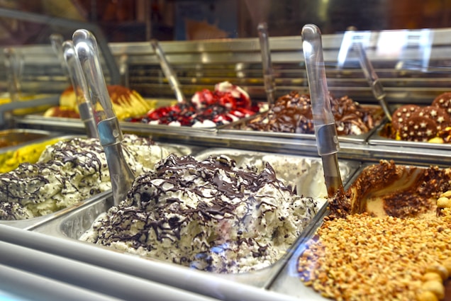 A display of various gelato flavors, with metal scoops positioned in each container. The gelato includes flavors with chocolate, nuts, and fruit toppings visible. The setting appears to be a gelato shop with multiple trays arranged in a row.
