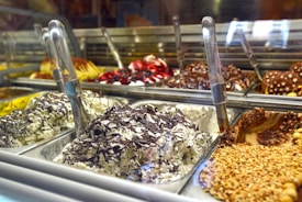 A display of various gelato flavors, with metal scoops positioned in each container. The gelato includes flavors with chocolate, nuts, and fruit toppings visible. The setting appears to be a gelato shop with multiple trays arranged in a row.