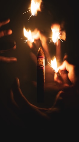 Several hands are holding lit lighters around a cigar, creating a dramatic and warm glow that illuminates the scene.