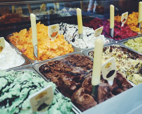 A variety of colorful gelato flavors are displayed in metal trays, each with a separate creamy texture and distinctive hue. Visible flavors include mint with chocolate drizzle, bright orange melon, vanilla with cookie bits, rich chocolate, and vibrant red berry. Each tray is marked with a flavor label and equipped with a serving spatula.