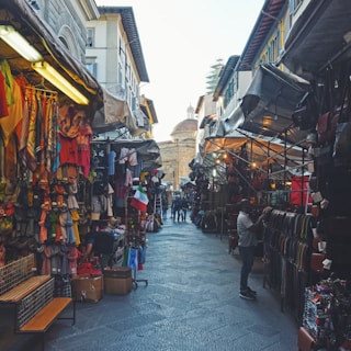 A bustling outdoor market with stalls on both sides offering various goods such as clothes, bags, and other colorful merchandise. The scene is set between narrow streets with historic buildings, and the sky is visible in the background. People are seen browsing and shopping.