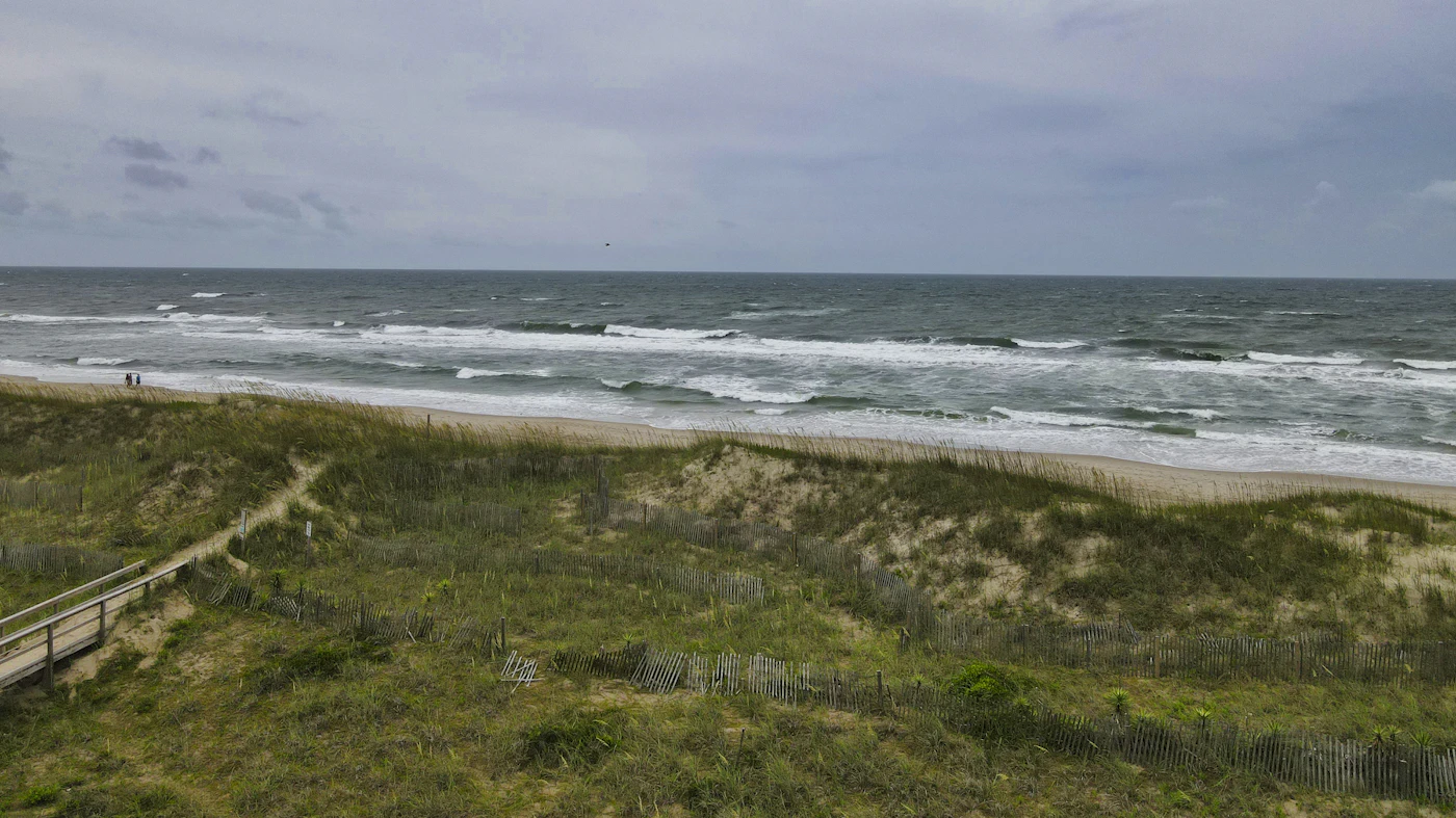 Grassy dunes with sand fencing and a wooden walkway above a windy Outer Banks beach under gray skies