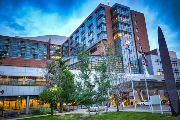 A large, multi-story modern building is prominently featured, with glass windows and a mix of brick and metallic elements. The foreground includes a few trees and a well-maintained lawn. On the building, a sculpture or logo with colorful elements is visible. Flags, including the United States flag, are positioned near the entrance where a white vehicle is parked.