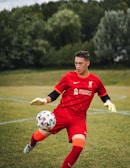 A soccer player wearing a red goalkeeper uniform is in action, attempting to control a soccer ball on a grassy field. The background is blurred, with green trees lining the field.