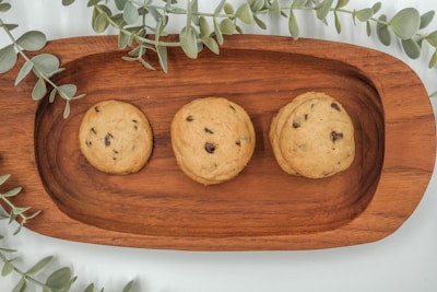 Three chocolate chip cookies are placed on a rectangular wooden tray. The cookies appear freshly baked with visible chocolate chips. Surrounding the tray are green eucalyptus leaves, adding a natural and fresh element to the composition.