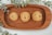 Three chocolate chip cookies are placed on a rectangular wooden tray. The cookies appear freshly baked with visible chocolate chips. Surrounding the tray are green eucalyptus leaves, adding a natural and fresh element to the composition.