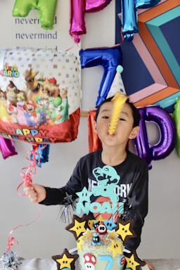 A child wearing a party hat is joyfully celebrating a birthday. They are holding a balloon themed with Super Mario characters. A colorful decorated cake with stars and various designs, including a '7' on it, is in front of the child. Background is adorned with balloons spelling out 'HAPPY' and numbers associated with birthday celebration.