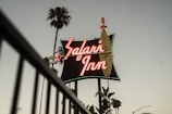 A vintage neon sign bearing the name 'Safari Inn' in bold, glowing letters is prominently displayed against a dusky sky. Tall palm trees flank the sign, adding a Californian or tropical ambiance to the scene. The overall atmosphere suggests a retro or nostalgic feel.