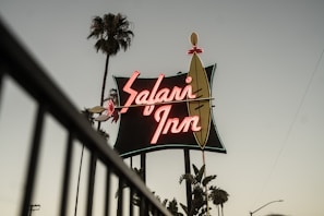 A vintage neon sign bearing the name 'Safari Inn' in bold, glowing letters is prominently displayed against a dusky sky. Tall palm trees flank the sign, adding a Californian or tropical ambiance to the scene. The overall atmosphere suggests a retro or nostalgic feel.