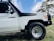 A white off-road vehicle with a visible snorkel and logo on the side. The image focuses on the front right section, including a rugged tire and a tree in the background.