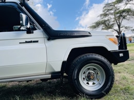 A white off-road vehicle with a visible snorkel and logo on the side. The image focuses on the front right section, including a rugged tire and a tree in the background.
