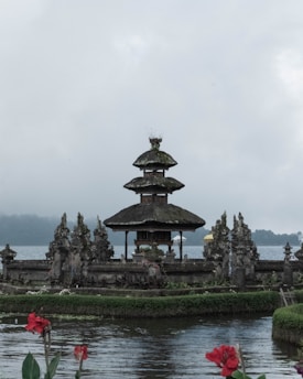 A traditional Balinese temple with a multi-tiered roof, surrounded by water and lush greenery. The temple is made of stone and features intricate carvings. In the foreground, red flowers bloom by the water's edge, adding a splash of color to the scene. The background shows a calm lake and distant mist-covered hills under an overcast sky.