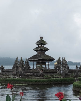 A traditional Balinese temple with a multi-tiered roof, surrounded by water and lush greenery. The temple is made of stone and features intricate carvings. In the foreground, red flowers bloom by the water's edge, adding a splash of color to the scene. The background shows a calm lake and distant mist-covered hills under an overcast sky.