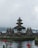 A traditional Balinese temple with a multi-tiered roof, surrounded by water and lush greenery. The temple is made of stone and features intricate carvings. In the foreground, red flowers bloom by the water's edge, adding a splash of color to the scene. The background shows a calm lake and distant mist-covered hills under an overcast sky.