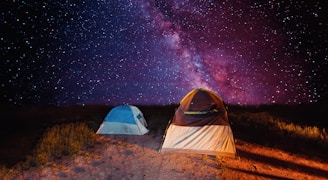 A stunning night sky filled with an array of stars, including the Milky Way, provides a breathtaking backdrop for two tents set up on a grassy patch. The larger tent is brown and white, while the smaller one is blue, both illuminated by a soft light, enhancing their presence against the vibrant cosmic display.
