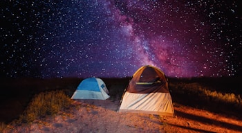 A stunning night sky filled with an array of stars, including the Milky Way, provides a breathtaking backdrop for two tents set up on a grassy patch. The larger tent is brown and white, while the smaller one is blue, both illuminated by a soft light, enhancing their presence against the vibrant cosmic display.