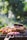 A barbecue grill loaded with a variety of meats and vegetables, including pieces of grilled chicken, ribs, and sliced red onions. The background features a blurred, lush green garden setting, creating a pleasant outdoor atmosphere.