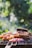 A barbecue grill loaded with a variety of meats and vegetables, including pieces of grilled chicken, ribs, and sliced red onions. The background features a blurred, lush green garden setting, creating a pleasant outdoor atmosphere.