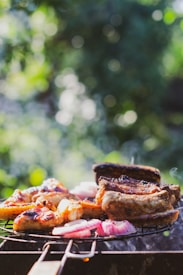 A barbecue grill loaded with a variety of meats and vegetables, including pieces of grilled chicken, ribs, and sliced red onions. The background features a blurred, lush green garden setting, creating a pleasant outdoor atmosphere.