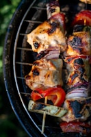 Grilled skewers featuring succulent pieces of meat, charred to perfection, interspersed with red bell peppers, slices of zucchini, and red onions. The skewers rest on a grill, with a vibrant outdoor ambiance.