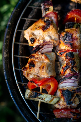 Grilled skewers featuring succulent pieces of meat, charred to perfection, interspersed with red bell peppers, slices of zucchini, and red onions. The skewers rest on a grill, with a vibrant outdoor ambiance.