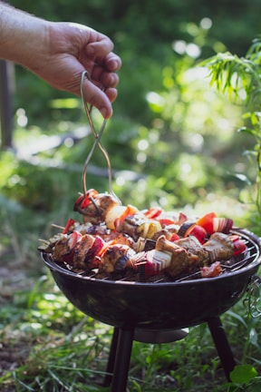 A hand holding metal tongs is seen lifting skewers filled with colorful vegetables and meat above a small black charcoal grill. The grill is set outdoors on a lush green grassy area, with sunlight filtering through the surrounding foliage.
