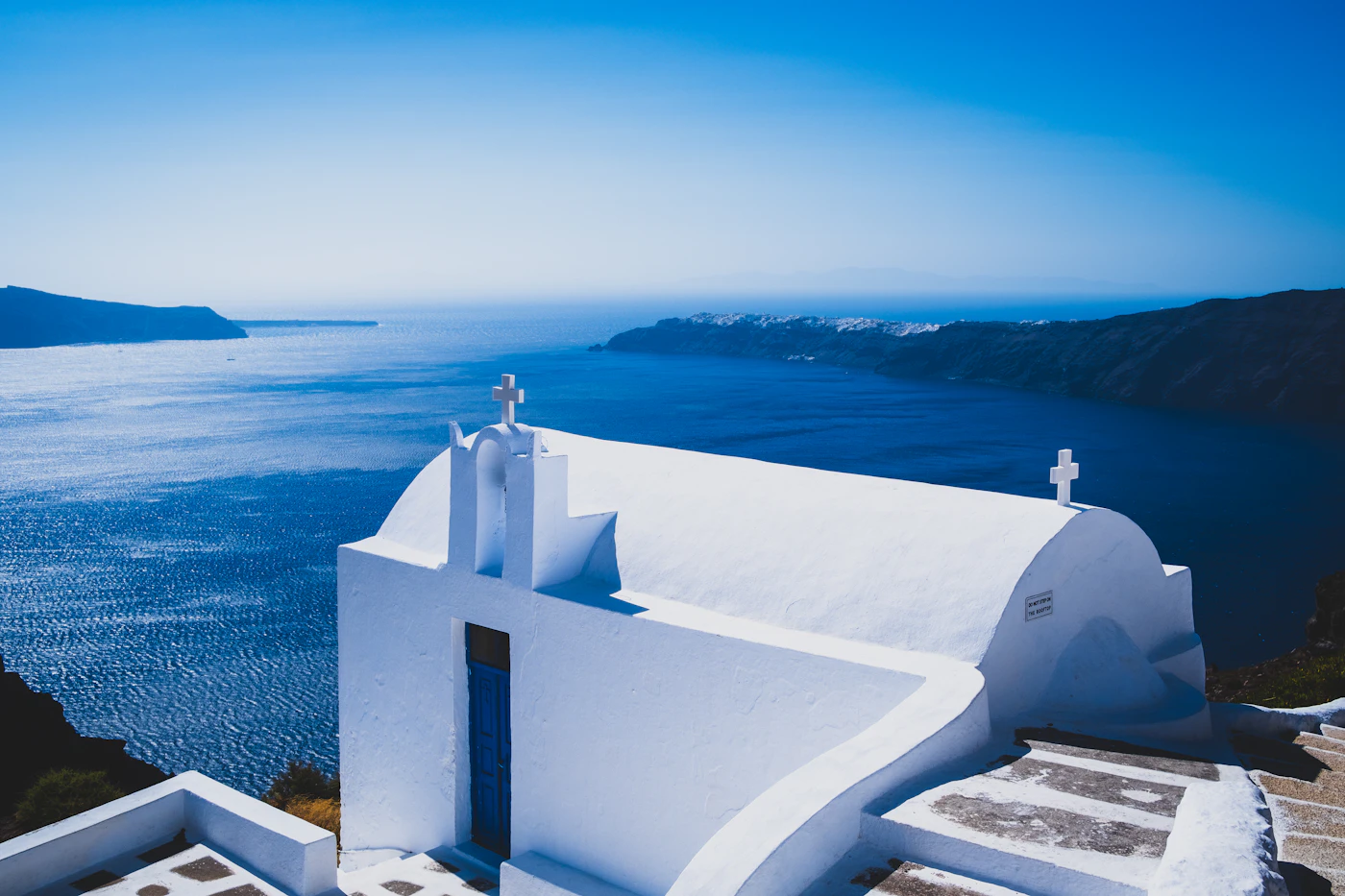 A small whitewashed chapel with two crosses on the caldera edge at Imerovigli, the sea and the far rim of the volcano behind it at dusk