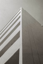 A monochrome photograph of a modern, geometric building taken from a low angle, emphasizing its height and angular design. The facade features a series of horizontal stripes and a grid pattern, with a minimalist and architectural aesthetic.