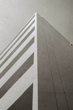 A monochrome photograph of a modern, geometric building taken from a low angle, emphasizing its height and angular design. The facade features a series of horizontal stripes and a grid pattern, with a minimalist and architectural aesthetic.