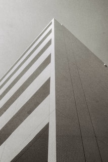 A monochrome photograph of a modern, geometric building taken from a low angle, emphasizing its height and angular design. The facade features a series of horizontal stripes and a grid pattern, with a minimalist and architectural aesthetic.