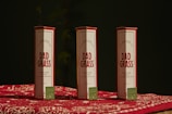 Three packages labeled 'Dad Grass' are placed upright on a surface covered with a red and white patterned cloth. Each package has a smaller, green box with text and a leaf logo on it.